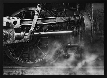 A dramatic, black and white close-up photograph of the wheels and driving rods of a steam locomotive. Puffs of steam rise from the tracks, partially obscuring the lower part of the machinery. Print