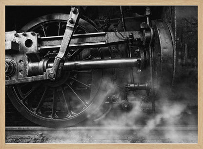 A dramatic, black and white close-up photograph of the wheels and driving rods of a steam locomotive. Puffs of steam rise from the tracks, partially obscuring the lower part of the machinery. Print