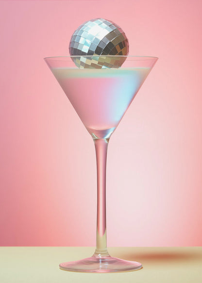 A sleek martini glass filled with an iridescent pink and blue cocktail is centered against a solid pink background. A small, shiny silver disco ball rests on the rim of the glass, completing the festive and stylish composition. Decor