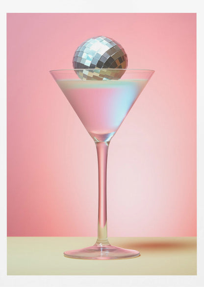A sleek martini glass filled with an iridescent pink and blue cocktail is centered against a solid pink background. A small, shiny silver disco ball rests on the rim of the glass, completing the festive and stylish composition. Decor