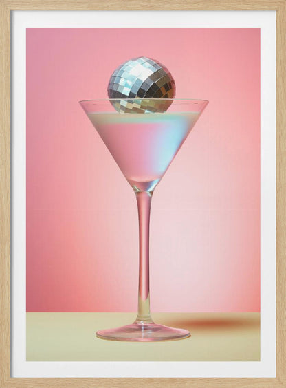 A sleek martini glass filled with an iridescent pink and blue cocktail is centered against a solid pink background. A small, shiny silver disco ball rests on the rim of the glass, completing the festive and stylish composition. Decor