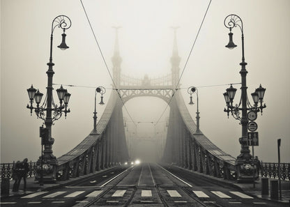 A framed, symmetrical black and white photograph of a large, ornate metal bridge disappearing into thick fog. Vintage lampposts stand on either side in the foreground, and the road and tram tracks lead the eye into the misty distance where faint car headlights glow. Print