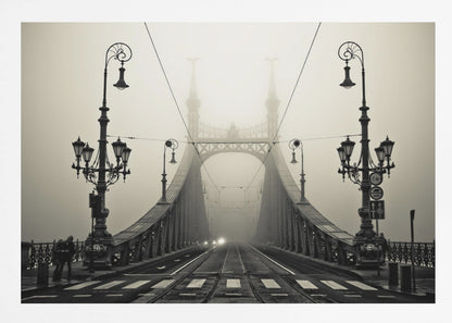 A framed, symmetrical black and white photograph of a large, ornate metal bridge disappearing into thick fog. Vintage lampposts stand on either side in the foreground, and the road and tram tracks lead the eye into the misty distance where faint car headlights glow. Print