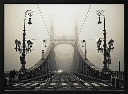 A framed, symmetrical black and white photograph of a large, ornate metal bridge disappearing into thick fog. Vintage lampposts stand on either side in the foreground, and the road and tram tracks lead the eye into the misty distance where faint car headlights glow. Print