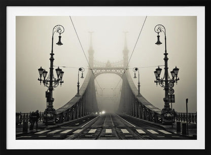 A framed, symmetrical black and white photograph of a large, ornate metal bridge disappearing into thick fog. Vintage lampposts stand on either side in the foreground, and the road and tram tracks lead the eye into the misty distance where faint car headlights glow. Print