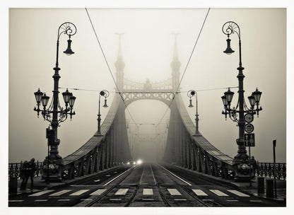 A framed, symmetrical black and white photograph of a large, ornate metal bridge disappearing into thick fog. Vintage lampposts stand on either side in the foreground, and the road and tram tracks lead the eye into the misty distance where faint car headlights glow. Print