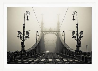 A framed, symmetrical black and white photograph of a large, ornate metal bridge disappearing into thick fog. Vintage lampposts stand on either side in the foreground, and the road and tram tracks lead the eye into the misty distance where faint car headlights glow. Print