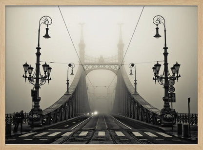 A framed, symmetrical black and white photograph of a large, ornate metal bridge disappearing into thick fog. Vintage lampposts stand on either side in the foreground, and the road and tram tracks lead the eye into the misty distance where faint car headlights glow. Print
