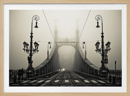 A framed, symmetrical black and white photograph of a large, ornate metal bridge disappearing into thick fog. Vintage lampposts stand on either side in the foreground, and the road and tram tracks lead the eye into the misty distance where faint car headlights glow. Print