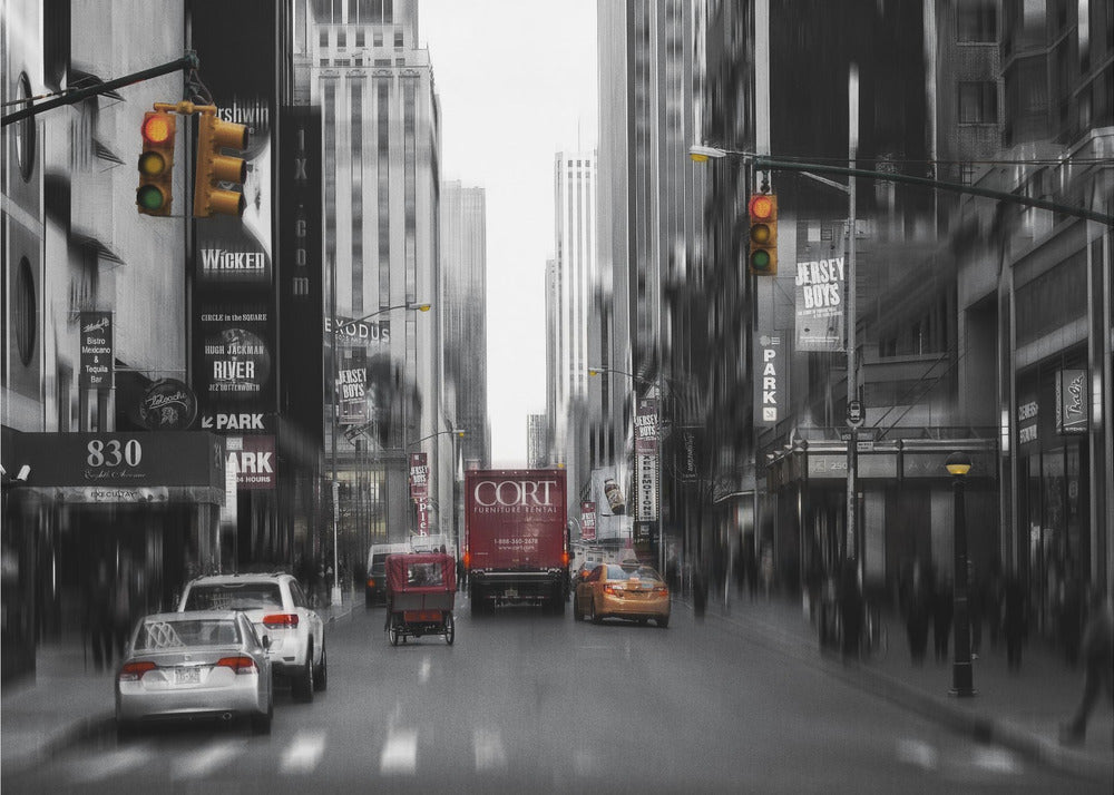 A framed, stylized photograph of a busy New York City street, presented mostly in black and white with a motion blur effect. Pops of color highlight red vehicles and yellow traffic lights, drawing focus down the urban canyon lined with skyscrapers and Broadway signs. Decor