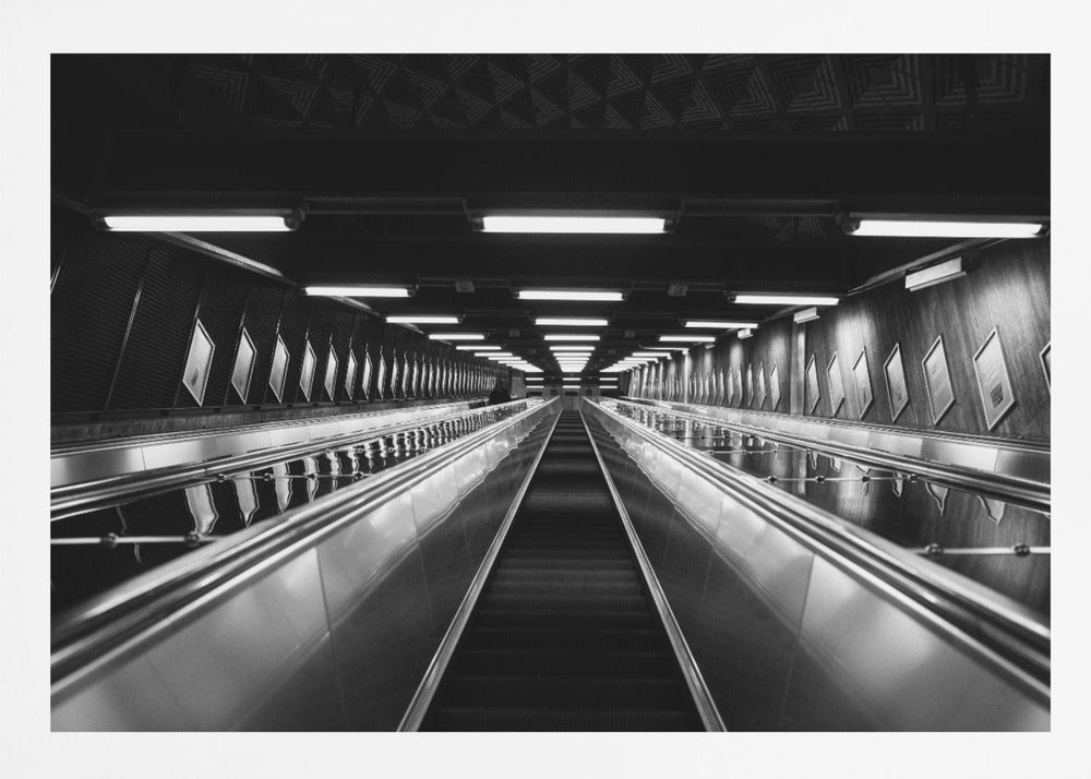 A framed, black and white, low-angle photograph looking up a long, steep, and empty escalator in a subway tunnel. The symmetrical composition uses the shiny metal handrails and rows of overhead lights as leading lines, creating a dramatic sense of depth and perspective. Print
