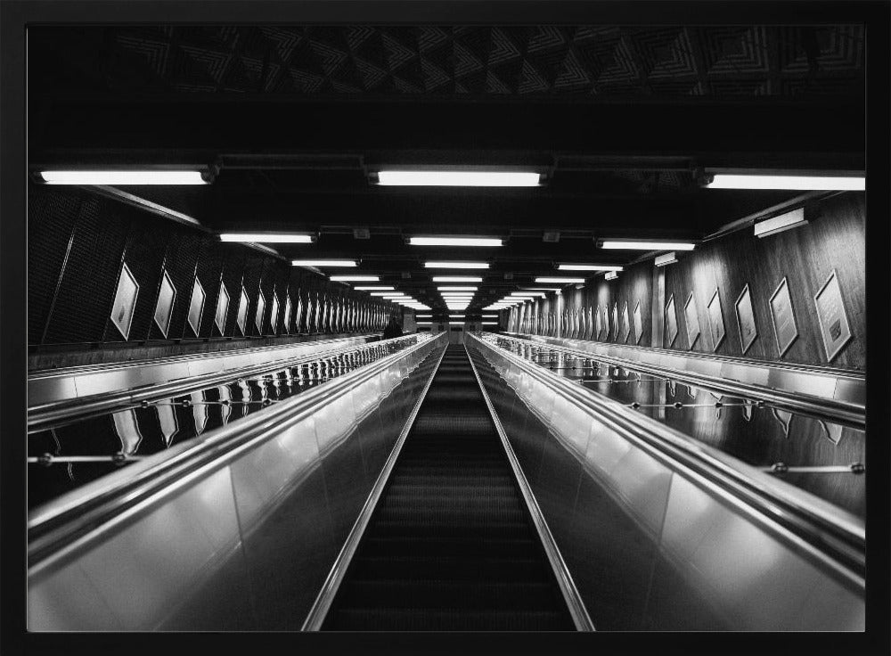 A framed, black and white, low-angle photograph looking up a long, steep, and empty escalator in a subway tunnel. The symmetrical composition uses the shiny metal handrails and rows of overhead lights as leading lines, creating a dramatic sense of depth and perspective. Print