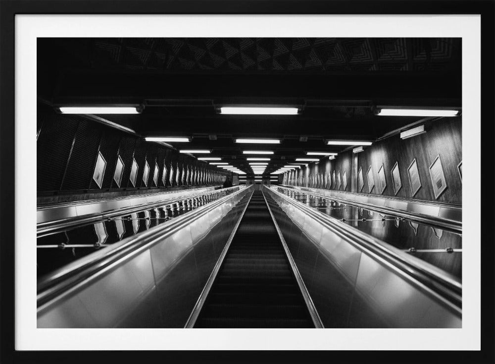 A framed, black and white, low-angle photograph looking up a long, steep, and empty escalator in a subway tunnel. The symmetrical composition uses the shiny metal handrails and rows of overhead lights as leading lines, creating a dramatic sense of depth and perspective. Print