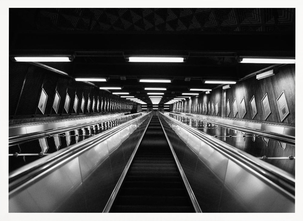 A framed, black and white, low-angle photograph looking up a long, steep, and empty escalator in a subway tunnel. The symmetrical composition uses the shiny metal handrails and rows of overhead lights as leading lines, creating a dramatic sense of depth and perspective. Print