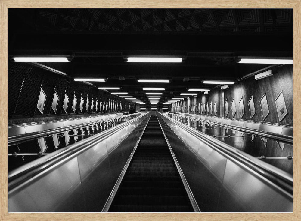 A framed, black and white, low-angle photograph looking up a long, steep, and empty escalator in a subway tunnel. The symmetrical composition uses the shiny metal handrails and rows of overhead lights as leading lines, creating a dramatic sense of depth and perspective. Print