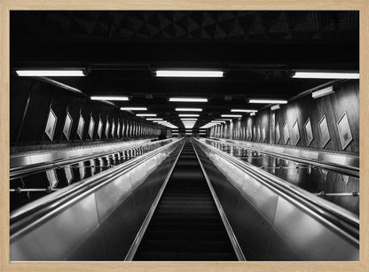 A framed, black and white, low-angle photograph looking up a long, steep, and empty escalator in a subway tunnel. The symmetrical composition uses the shiny metal handrails and rows of overhead lights as leading lines, creating a dramatic sense of depth and perspective. Print