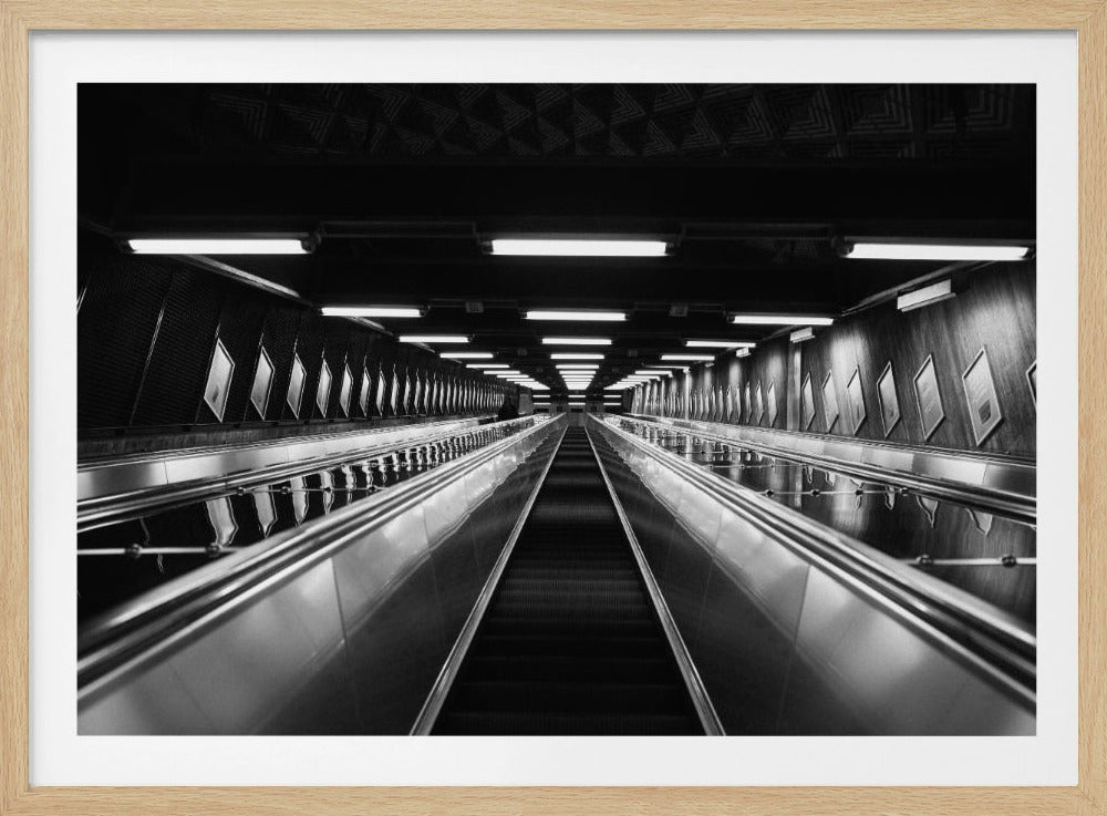 A framed, black and white, low-angle photograph looking up a long, steep, and empty escalator in a subway tunnel. The symmetrical composition uses the shiny metal handrails and rows of overhead lights as leading lines, creating a dramatic sense of depth and perspective. Print
