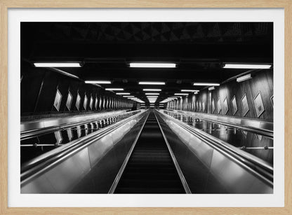 A framed, black and white, low-angle photograph looking up a long, steep, and empty escalator in a subway tunnel. The symmetrical composition uses the shiny metal handrails and rows of overhead lights as leading lines, creating a dramatic sense of depth and perspective. Print