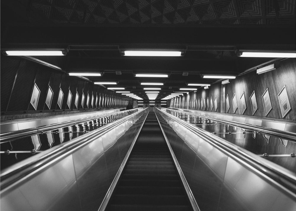 A framed, black and white, low-angle photograph looking up a long, steep, and empty escalator in a subway tunnel. The symmetrical composition uses the shiny metal handrails and rows of overhead lights as leading lines, creating a dramatic sense of depth and perspective. Print