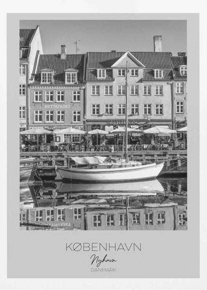 A black and white framed poster of Nyhavn in Copenhagen. The photograph features classic Danish buildings lining a canal, with a sailboat moored in the water. The buildings and boat are perfectly reflected in the still water. At the bottom, text reads 'KØBENHAVN', 'Nyhavn', and 'DANMARK'. Wall Art