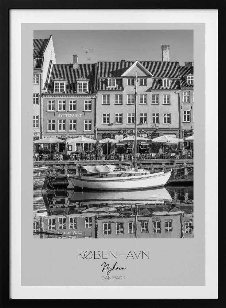 A black and white framed poster of Nyhavn in Copenhagen. The photograph features classic Danish buildings lining a canal, with a sailboat moored in the water. The buildings and boat are perfectly reflected in the still water. At the bottom, text reads 'KØBENHAVN', 'Nyhavn', and 'DANMARK'. Wall Art