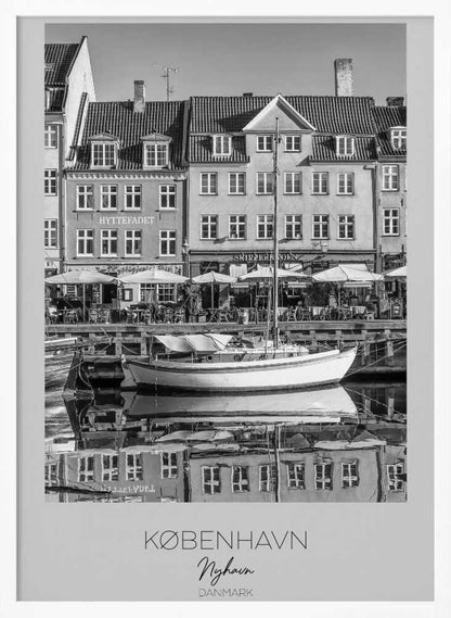 A black and white framed poster of Nyhavn in Copenhagen. The photograph features classic Danish buildings lining a canal, with a sailboat moored in the water. The buildings and boat are perfectly reflected in the still water. At the bottom, text reads 'KØBENHAVN', 'Nyhavn', and 'DANMARK'. Wall Art