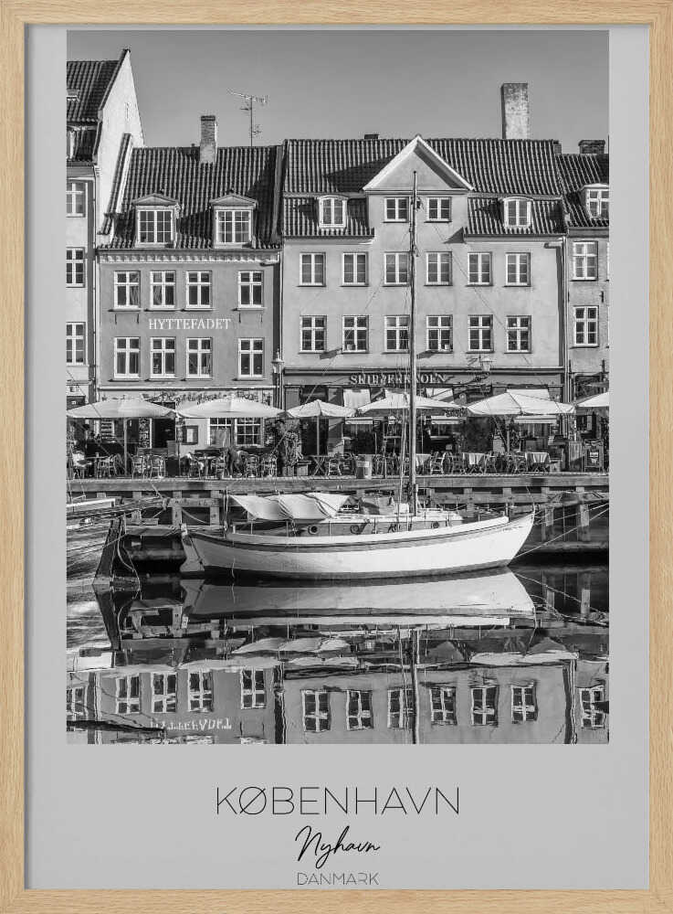 A black and white framed poster of Nyhavn in Copenhagen. The photograph features classic Danish buildings lining a canal, with a sailboat moored in the water. The buildings and boat are perfectly reflected in the still water. At the bottom, text reads 'KØBENHAVN', 'Nyhavn', and 'DANMARK'. Wall Art