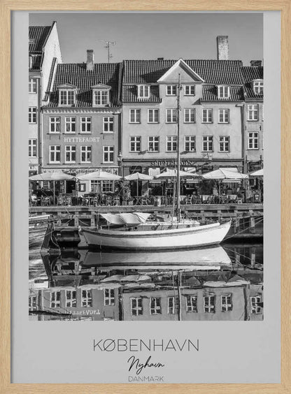 A black and white framed poster of Nyhavn in Copenhagen. The photograph features classic Danish buildings lining a canal, with a sailboat moored in the water. The buildings and boat are perfectly reflected in the still water. At the bottom, text reads 'KØBENHAVN', 'Nyhavn', and 'DANMARK'. Wall Art