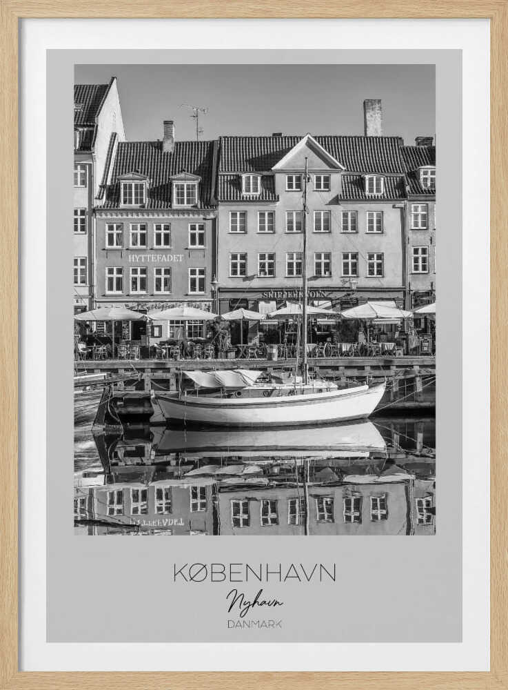 A black and white framed poster of Nyhavn in Copenhagen. The photograph features classic Danish buildings lining a canal, with a sailboat moored in the water. The buildings and boat are perfectly reflected in the still water. At the bottom, text reads 'KØBENHAVN', 'Nyhavn', and 'DANMARK'. Wall Art