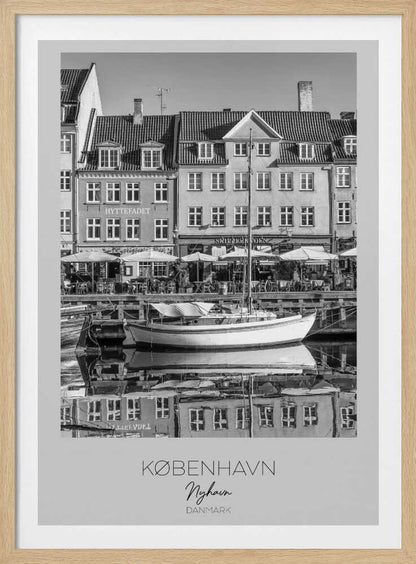A black and white framed poster of Nyhavn in Copenhagen. The photograph features classic Danish buildings lining a canal, with a sailboat moored in the water. The buildings and boat are perfectly reflected in the still water. At the bottom, text reads 'KØBENHAVN', 'Nyhavn', and 'DANMARK'. Wall Art