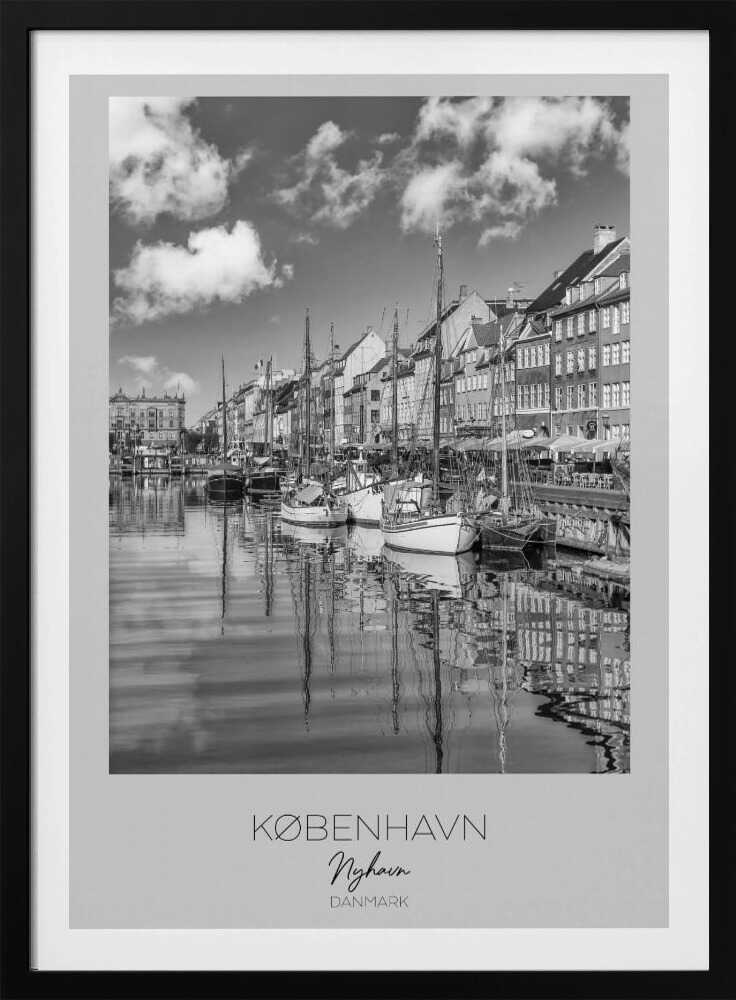 A black and white poster of Nyhavn harbor in Copenhagen. The photograph shows traditional sailboats and ships moored along a canal, with a line of historic, multi-story buildings on the far bank. The sky has fluffy clouds, and the entire scene is reflected in the calm water. The poster has a white border with the text 'KØBENHAVN', 'Nyhavn', and 'DANMARK' at the bottom, and is shown within a black frame. Poster