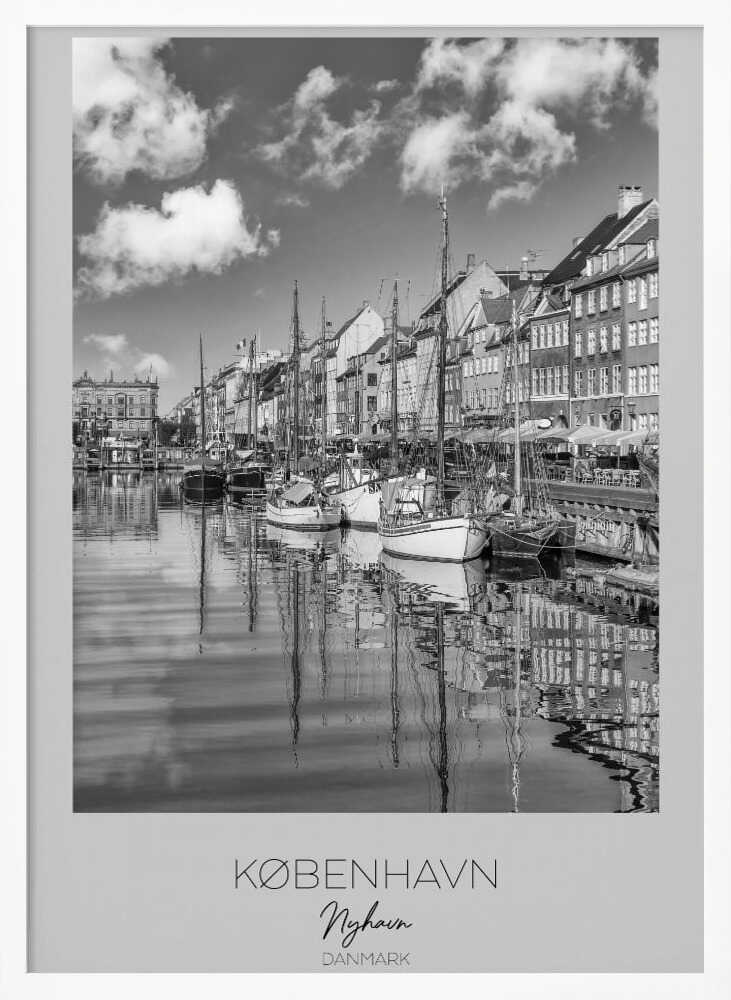 A black and white poster of Nyhavn harbor in Copenhagen. The photograph shows traditional sailboats and ships moored along a canal, with a line of historic, multi-story buildings on the far bank. The sky has fluffy clouds, and the entire scene is reflected in the calm water. The poster has a white border with the text 'KØBENHAVN', 'Nyhavn', and 'DANMARK' at the bottom, and is shown within a black frame. Poster