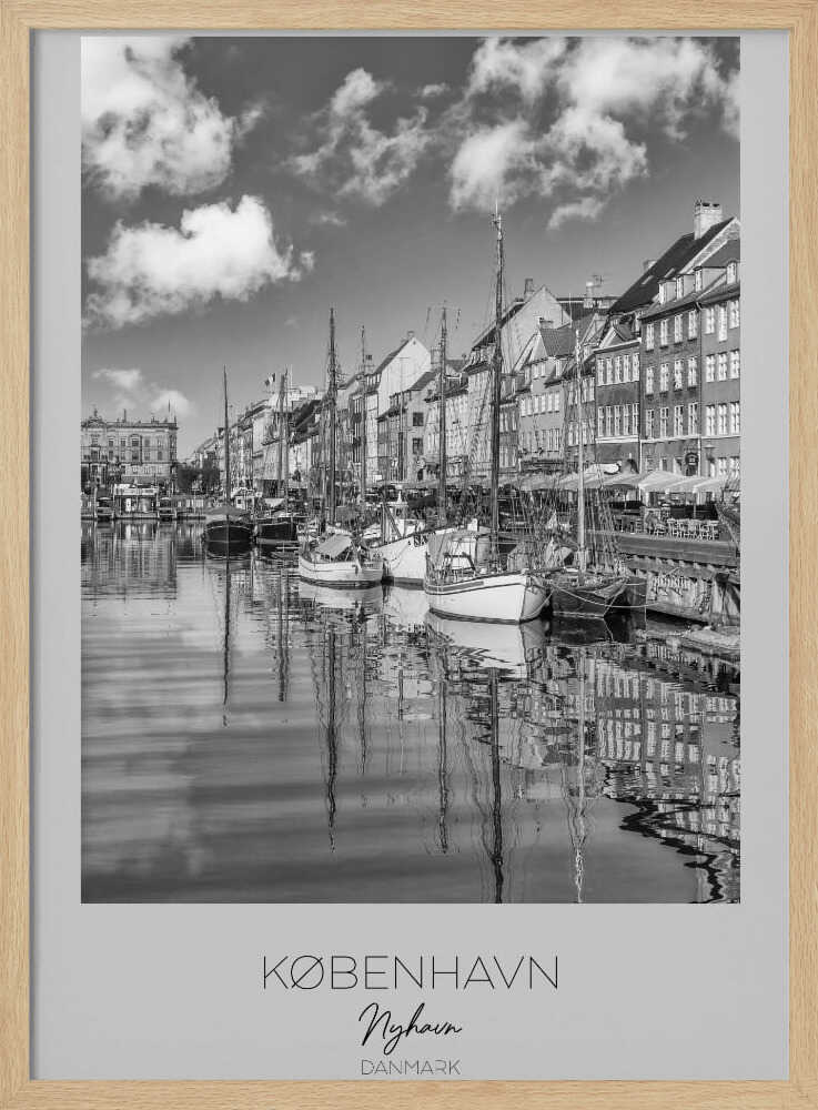 A black and white poster of Nyhavn harbor in Copenhagen. The photograph shows traditional sailboats and ships moored along a canal, with a line of historic, multi-story buildings on the far bank. The sky has fluffy clouds, and the entire scene is reflected in the calm water. The poster has a white border with the text 'KØBENHAVN', 'Nyhavn', and 'DANMARK' at the bottom, and is shown within a black frame. Poster