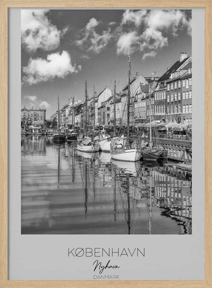 A black and white poster of Nyhavn harbor in Copenhagen. The photograph shows traditional sailboats and ships moored along a canal, with a line of historic, multi-story buildings on the far bank. The sky has fluffy clouds, and the entire scene is reflected in the calm water. The poster has a white border with the text 'KØBENHAVN', 'Nyhavn', and 'DANMARK' at the bottom, and is shown within a black frame. Poster