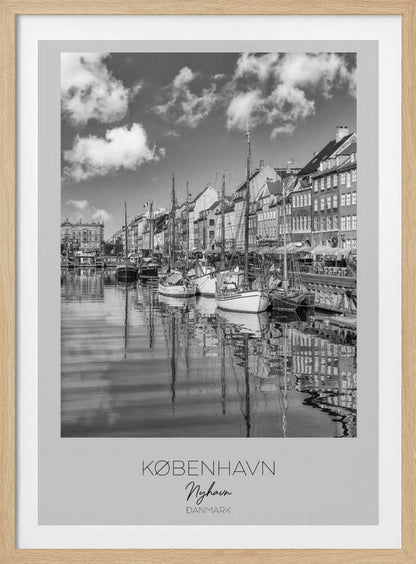 A black and white poster of Nyhavn harbor in Copenhagen. The photograph shows traditional sailboats and ships moored along a canal, with a line of historic, multi-story buildings on the far bank. The sky has fluffy clouds, and the entire scene is reflected in the calm water. The poster has a white border with the text 'KØBENHAVN', 'Nyhavn', and 'DANMARK' at the bottom, and is shown within a black frame. Poster