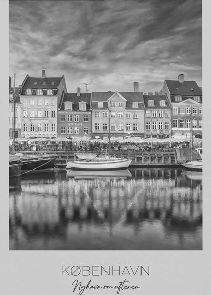 A framed black and white photograph of the Nyhavn harbor in Copenhagen, Denmark. The image shows a row of classic gabled houses along the waterfront, with several sailboats moored in the canal. The buildings and a dramatic cloudy sky are reflected in the water. At the bottom, a white border contains the text 'KØBENHAVN', 'Nyhavn om aftenen', and 'DANMARK'. Wall Art