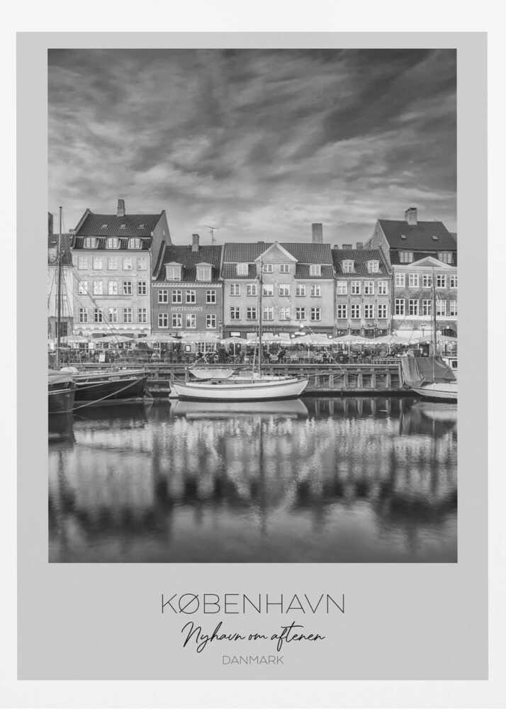 A framed black and white photograph of the Nyhavn harbor in Copenhagen, Denmark. The image shows a row of classic gabled houses along the waterfront, with several sailboats moored in the canal. The buildings and a dramatic cloudy sky are reflected in the water. At the bottom, a white border contains the text 'KØBENHAVN', 'Nyhavn om aftenen', and 'DANMARK'. Wall Art