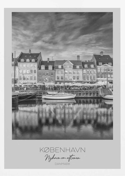 A framed black and white photograph of the Nyhavn harbor in Copenhagen, Denmark. The image shows a row of classic gabled houses along the waterfront, with several sailboats moored in the canal. The buildings and a dramatic cloudy sky are reflected in the water. At the bottom, a white border contains the text 'KØBENHAVN', 'Nyhavn om aftenen', and 'DANMARK'. Wall Art