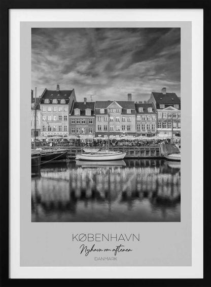 A framed black and white photograph of the Nyhavn harbor in Copenhagen, Denmark. The image shows a row of classic gabled houses along the waterfront, with several sailboats moored in the canal. The buildings and a dramatic cloudy sky are reflected in the water. At the bottom, a white border contains the text 'KØBENHAVN', 'Nyhavn om aftenen', and 'DANMARK'. Wall Art