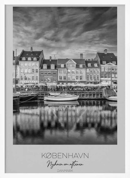 A framed black and white photograph of the Nyhavn harbor in Copenhagen, Denmark. The image shows a row of classic gabled houses along the waterfront, with several sailboats moored in the canal. The buildings and a dramatic cloudy sky are reflected in the water. At the bottom, a white border contains the text 'KØBENHAVN', 'Nyhavn om aftenen', and 'DANMARK'. Wall Art
