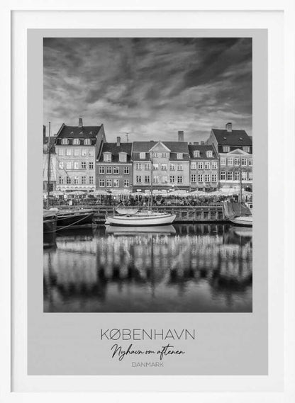 A framed black and white photograph of the Nyhavn harbor in Copenhagen, Denmark. The image shows a row of classic gabled houses along the waterfront, with several sailboats moored in the canal. The buildings and a dramatic cloudy sky are reflected in the water. At the bottom, a white border contains the text 'KØBENHAVN', 'Nyhavn om aftenen', and 'DANMARK'. Wall Art