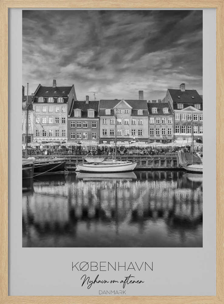 A framed black and white photograph of the Nyhavn harbor in Copenhagen, Denmark. The image shows a row of classic gabled houses along the waterfront, with several sailboats moored in the canal. The buildings and a dramatic cloudy sky are reflected in the water. At the bottom, a white border contains the text 'KØBENHAVN', 'Nyhavn om aftenen', and 'DANMARK'. Wall Art
