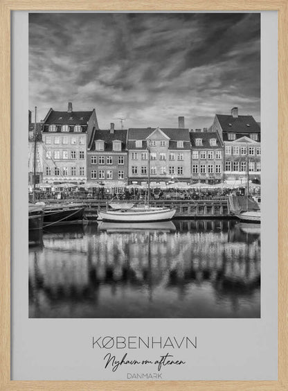 A framed black and white photograph of the Nyhavn harbor in Copenhagen, Denmark. The image shows a row of classic gabled houses along the waterfront, with several sailboats moored in the canal. The buildings and a dramatic cloudy sky are reflected in the water. At the bottom, a white border contains the text 'KØBENHAVN', 'Nyhavn om aftenen', and 'DANMARK'. Wall Art