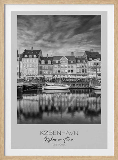 A framed black and white photograph of the Nyhavn harbor in Copenhagen, Denmark. The image shows a row of classic gabled houses along the waterfront, with several sailboats moored in the canal. The buildings and a dramatic cloudy sky are reflected in the water. At the bottom, a white border contains the text 'KØBENHAVN', 'Nyhavn om aftenen', and 'DANMARK'. Wall Art
