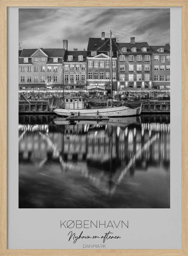 A black and white poster of Nyhavn harbor in Copenhagen at night. Historic buildings with glowing windows line the canal, and their lights create shimmering reflections in the water. A boat is docked in the foreground. The poster has text at the bottom that reads 'KØBENHAVN, Nyhavn om aftenen, DANMARK'. Poster