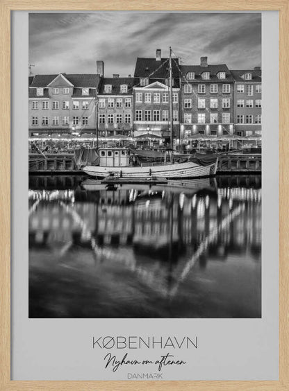 A black and white poster of Nyhavn harbor in Copenhagen at night. Historic buildings with glowing windows line the canal, and their lights create shimmering reflections in the water. A boat is docked in the foreground. The poster has text at the bottom that reads 'KØBENHAVN, Nyhavn om aftenen, DANMARK'. Poster