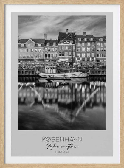 A black and white poster of Nyhavn harbor in Copenhagen at night. Historic buildings with glowing windows line the canal, and their lights create shimmering reflections in the water. A boat is docked in the foreground. The poster has text at the bottom that reads 'KØBENHAVN, Nyhavn om aftenen, DANMARK'. Poster