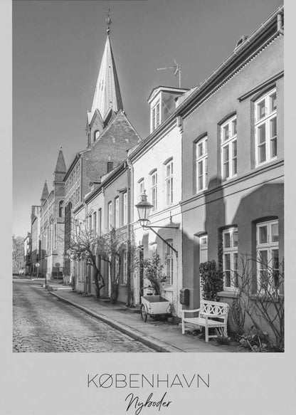 A black and white framed travel poster of a charming cobblestone street in Nyboder, Copenhagen. The photograph shows a sunlit row of historic houses with a prominent church steeple in the background. At the bottom of the poster, the words 'KØBENHAVN', 'Nyboder', and 'DANMARK' are written. Wall Art
