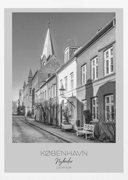 A black and white framed travel poster of a charming cobblestone street in Nyboder, Copenhagen. The photograph shows a sunlit row of historic houses with a prominent church steeple in the background. At the bottom of the poster, the words 'KØBENHAVN', 'Nyboder', and 'DANMARK' are written. Wall Art