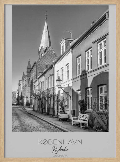 A black and white framed travel poster of a charming cobblestone street in Nyboder, Copenhagen. The photograph shows a sunlit row of historic houses with a prominent church steeple in the background. At the bottom of the poster, the words 'KØBENHAVN', 'Nyboder', and 'DANMARK' are written. Wall Art